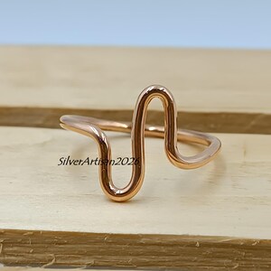 Può includere: Un anello in oro rosa dal design astratto unico. L'anello presenta una linea ondulata continua che forma una serie di anelli e curve. L'anello è mostrato su una superficie di legno. Il testo "SilverArtisan2020" è visibile.