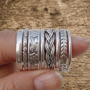 May include: A set of eight silver-toned rings featuring various designs including a smooth band, a geometric pattern, a floral motif, a braided texture, and a leaf-like pattern. The rings are arranged side-by-side, showcasing different widths and finishes.