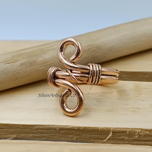 K&ouml;nnte beinhalten: Ein kupferfarbener Ring mit einem einzigartigen, handgefertigten Design. Der Ring hat eine gewickelte Ober- und Unterseite, die durch ein gewickeltes Band verbunden sind. Der Ring wird auf einer Holzoberfl&auml;che pr&auml;sentiert.