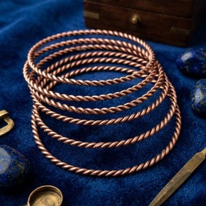 May include: A set of five circular copper-toned bangles featuring a twisted rope-like texture. The bracelets are arranged in a stacked formation, showcasing the metallic finish and the consistent spiral pattern of each individual bangle band.
