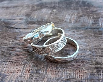 May include: Three silver rings stacked together. The top ring has a hammered texture, the middle ring features an embossed floral pattern, and the bottom ring has a hammered finish. The text SilverArtisan2026 is visible on the top ring.