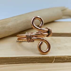 K&ouml;nnte beinhalten: Ein handgefertigter Kupferring mit einem einzigartigen, k&uuml;nstlerischen Design. Der Ring weist gewickelte und geschlungene Elemente auf, mit einem Band, das mit Kupferdraht umwickelt ist. Der Ring wird auf einer Holzoberfl&auml;che pr&auml;sentiert und zeigt seine komplizierten Details und seine warme, metallische Farbe.
