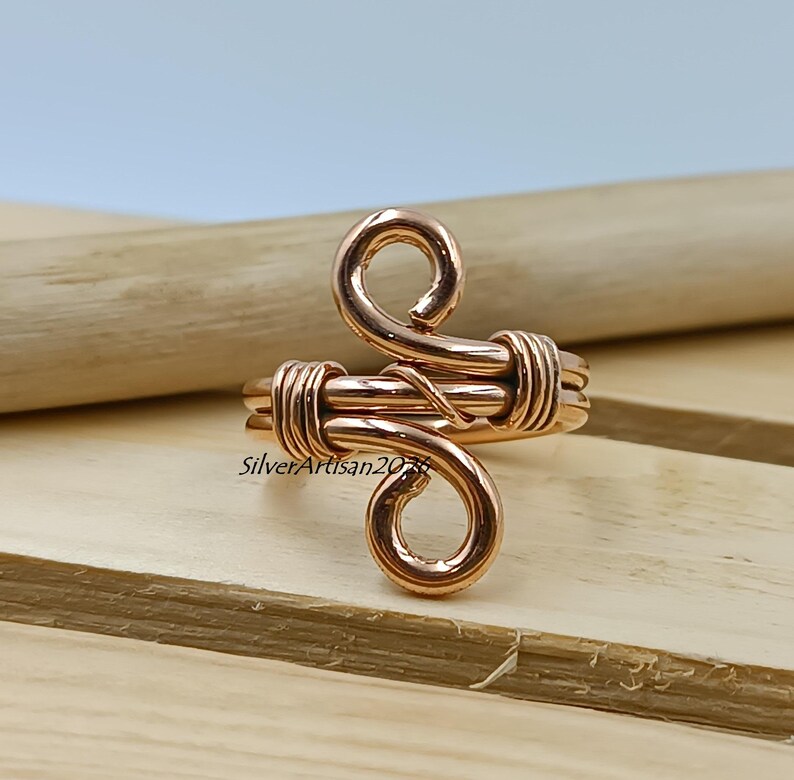 K&ouml;nnte beinhalten: Ein kupferfarbener Drahtring mit einem einzigartigen, wirbelnden Design. Der Ring hat einen zentralen gewickelten Abschnitt und zwei gebogene, offen endende Spiralen. Der Ring wird auf einer Holzoberfl&auml;che pr&auml;sentiert.