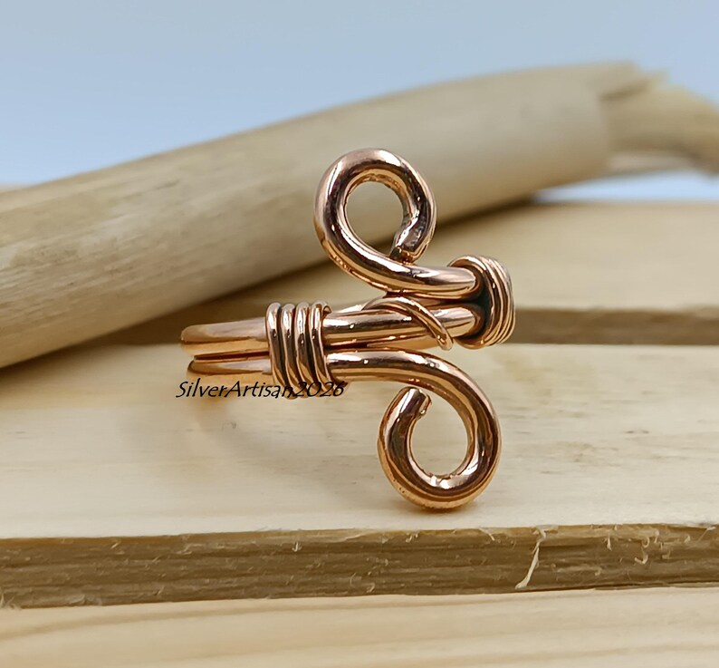 K&ouml;nnte beinhalten: Ein handgefertigter Kupferring mit einem einzigartigen, k&uuml;nstlerischen Design. Der Ring hat eine gewickelte Oberseite und ein gewickeltes Band, das eine Mischung aus Kurven und geraden Linien zeigt. Der Ring wird auf einer Holzoberfl&auml;che pr&auml;sentiert.