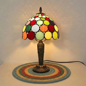 May include: A stained glass table lamp with a domed shade. The shade is decorated with colourful circular shapes in red, green, yellow, orange, and white. The lamp has a bronze base and sits on a round, multi-coloured rug.