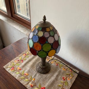 May include: A table lamp featuring a dome-shaped shade constructed from numerous circular stained glass pieces in various colours including red, green, blue, yellow, and white, set in a dark metal frame with a matching ornate metal base.