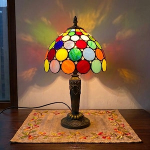 May include: A table lamp featuring a dome-shaped shade constructed from numerous circular glass pieces in vibrant colors including red, blue, green, orange, and white. The lamp has a dark metal base with an ornate, textured stem and a black power cord.