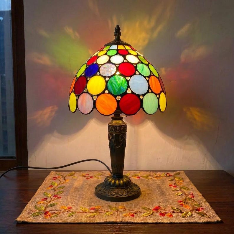 May include: A table lamp featuring a dome-shaped shade constructed from numerous circular glass pieces in vibrant colours including red, blue, green, orange, and white. The lamp has a dark metal base with an ornate, textured stem and a black power cord.