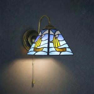 May include: A stained glass wall sconce with a nautical theme. The lamp has a triangular shade with a blue sky and sea design, and two yellow sailboats. The fixture has a brass finish and a pull chain.