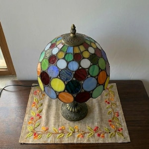 May include: A spherical stained glass table lamp featuring a multi-coloured pattern of circular glass pieces in shades of red, blue, green, yellow, and orange. The lamp has a dark metal base and a matching metal cap with a decorative perforated design.