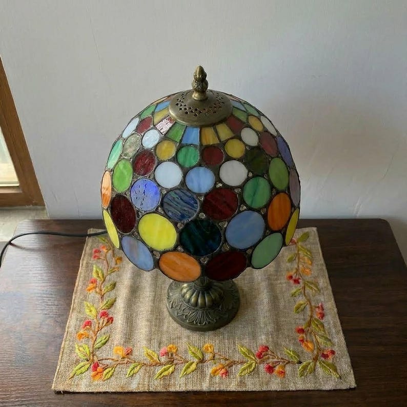 May include: A spherical stained glass table lamp featuring a multi-coloured pattern of circular glass pieces in shades of red, blue, green, yellow, and orange. The lamp has a dark metal base and a matching metal cap with a decorative perforated design.