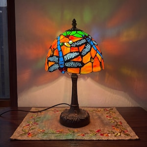 May include: A stained glass table lamp featuring a dome-shaped shade with a vibrant dragonfly motif in blue, orange, and green. The lamp has a dark metal base and emits a warm, colorful glow that casts patterned light onto the surrounding area.