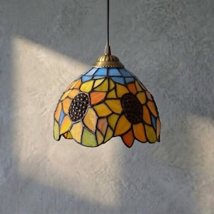 May include: A stained glass pendant light with a sunflower design. The lamp has a blue top, yellow and orange petals, and two dark brown centres. The fixture is suspended by a black cord and has a brass-coloured cap.