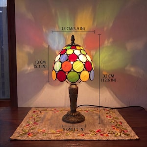 May include: A stained glass table lamp with a dome-shaped shade featuring multicoloured circular glass pieces in red, green, blue, and yellow. The lamp has a dark metal base and stands 32 cm tall, with a 15 cm wide shade and a 9 cm wide base.