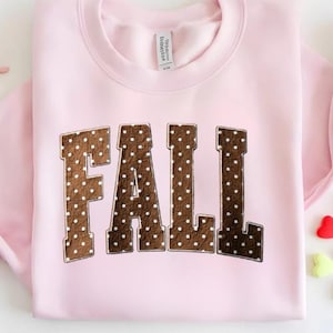 Könnte beinhalten: Ein hellrosa Sweatshirt mit Rundhalsausschnitt und dem Wort FALL in großen, braunen Blockbuchstaben. Jeder Buchstabe hat ein weißes Punktemuster und eine dünne weiße Umrandung. Das Kleidungsstück hat einen gerippten Ausschnitt und ein weißes Etikett.