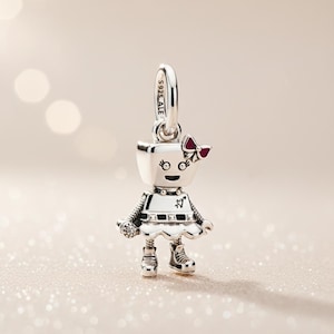 May include: A silver robot charm with a red bow, a white dress, and black boots. The charm has a loop at the top for a bracelet or necklace. The robot has a smiling face and a small heart design on its dress.