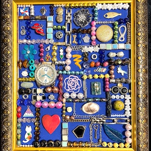以下が含まれることがあります： 青色の背景に額装されたコラージュで、様々なジュエリーや装飾品が特徴です。時計、ハート、猫、金、銀、カラフルな色合いの様々なビーズやチャームが含まれています。フレームは装飾的な金です。