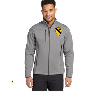 Op de afbeelding: Een grijze softshell jas met een volledige ritssluiting en een opstaande kraag. De jas heeft een zwart en geel embleem op de borst. De jas heeft twee zakken met ritssluiting aan de voorkant en één op de linkermouw.