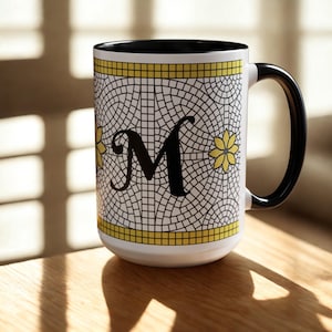 May include: White ceramic mug with a black handle and interior. The mug features a mosaic-style design with a large black letter "M" and yellow floral accents. A yellow border accents the top and bottom.