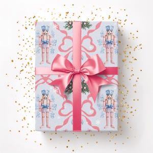Puede incluir: Un regalo envuelto con papel de regalo azul claro con un patrón de cascanueces rosas y copos de nieve. Una cinta y un lazo rosas están atados alrededor del regalo. Brillo dorado esparcido alrededor del regalo.