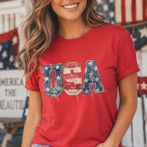 Könnte beinhalten: Ein rotes Kurzarm-T-Shirt mit dem Schriftzug USA auf der Brust. Die Buchstaben haben eine abgenutzte Jeans-Optik mit weißen Sternen sowie roten und weißen Streifen und sind an den Rändern mit sichtbaren, ausgefransten Nähten versehen.