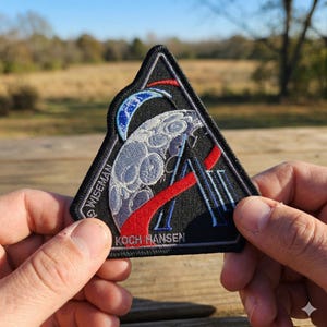 May include: A black, triangular patch featuring a detailed design of a moon, a crescent, and the letters "A" and "II". The patch includes the names "WISEMAN" and "KOCH HANSEN". The design incorporates red, white, and blue accents.