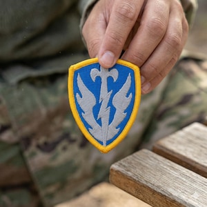 May include: A blue and gray military patch with a yellow border. The patch features a stylized lightning bolt and wings design. The patch is held up against a camouflage background.