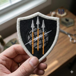 May include: A black and white embroidered patch with a shield shape. The patch features three spears, a globe, and a lightning bolt design. The border is white, and the patch is held in a hand.
