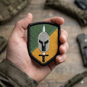 May include: A shield-shaped patch with a black border, featuring a Spartan helmet and spear design. The patch is divided diagonally, with green and yellow sections. The helmet is silver, and the spear is white.
