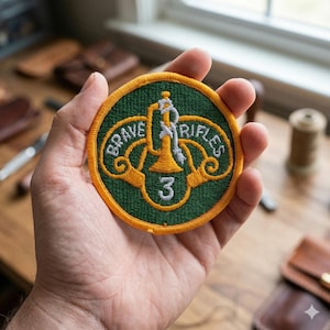 May include: A round, embroidered patch with a green background and a gold border. The patch features the words "BRAVE RIFLES" in a curved design, with a central emblem and the number "3". The emblem is a gold bugle with a white center.