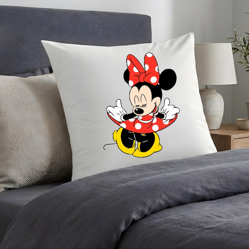 Puede incluir: Coj&iacute;n cuadrado blanco con un dise&ntilde;o de dibujos animados de Minnie Mouse. Minnie Mouse est&aacute; representada con un vestido rojo de lunares y un lazo, orejas negras y zapatos amarillos. El coj&iacute;n est&aacute; sobre una cama con ropa de cama gris.
