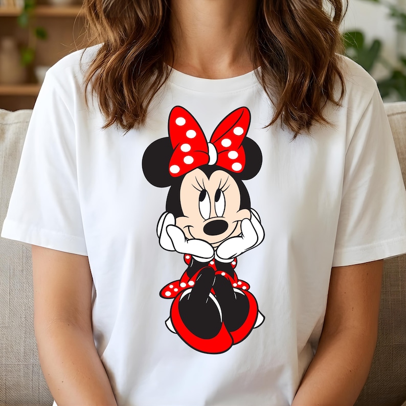 Puede incluir: Camiseta blanca con una ilustraci&oacute;n de dibujos animados de Minnie Mouse. Minnie Mouse tiene un lazo rojo con lunares blancos, un vestido rojo y negro y guantes blancos. El personaje sonr&iacute;e y apoya la barbilla en sus manos.