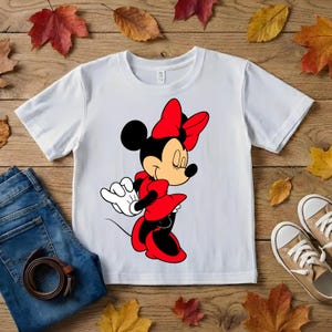 Puede incluir: Camiseta blanca con una ilustraci&oacute;n de dibujos animados de Minnie Mouse con un vestido y lazo rojos. El dise&ntilde;o est&aacute; centrado en la camiseta. La imagen tambi&eacute;n incluye vaqueros azules, un cintur&oacute;n marr&oacute;n y zapatillas de deporte color beige sobre una superficie de madera.