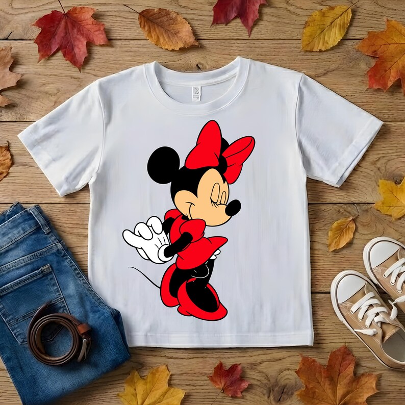 Puede incluir: Camiseta blanca con una ilustraci&oacute;n de dibujos animados de Minnie Mouse con un vestido y lazo rojos. El dise&ntilde;o est&aacute; centrado en la camiseta. La imagen tambi&eacute;n incluye vaqueros azules, un cintur&oacute;n marr&oacute;n y zapatillas de deporte color beige sobre una superficie de madera.