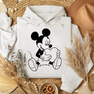 May include: White hoodie featuring a black and white illustration of Mickey Mouse holding a heart. The cartoon character is seated, with a smiling expression. The hoodie has a drawstring hood and a kangaroo pocket.