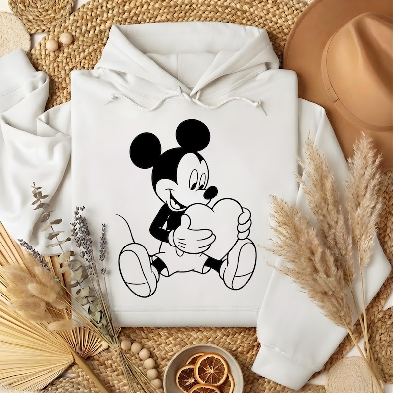 May include: White hoodie featuring a black and white illustration of Mickey Mouse holding a heart. The cartoon character is seated, with a smiling expression. The hoodie has a drawstring hood and a kangaroo pocket.