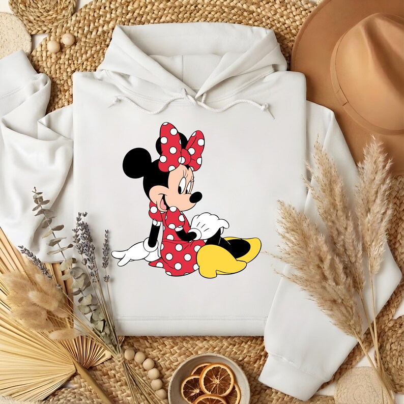 Puede incluir: Sudadera con capucha blanca con un gr&aacute;fico de Minnie Mouse. El gr&aacute;fico muestra a Minnie Mouse con un vestido de lunares rojos, un lazo a juego y zapatos amarillos. La sudadera tiene una capucha con cord&oacute;n y un bolsillo tipo canguro.