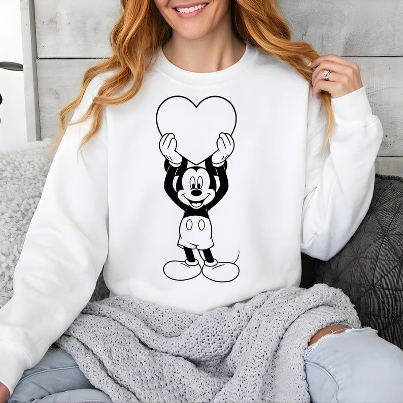 May include: White sweatshirt featuring a black and white graphic of a cartoon character holding a heart. The character has black ears, a black face, and white shorts. The heart is white. The sweatshirt has long sleeves.