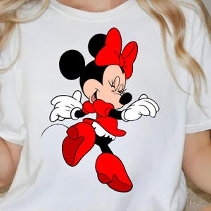 Puede incluir: Camiseta blanca con una ilustraci&oacute;n de dibujos animados de Minnie Mouse. Minnie Mouse lleva un vestido rojo, zapatos rojos y un lazo rojo. Sus orejas negras y su pelo negro son visibles. Sonr&iacute;e con los ojos cerrados y sus guantes blancos son visibles.