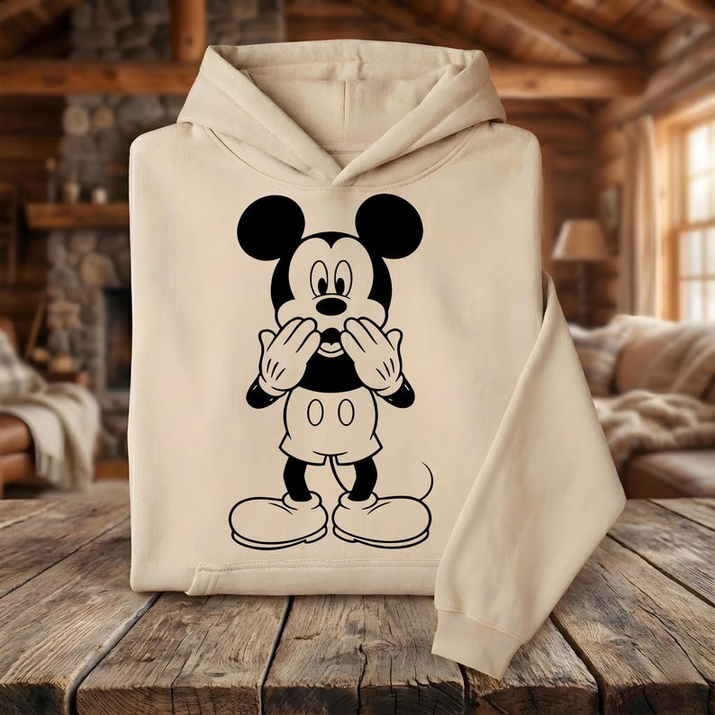 May include: A beige hoodie featuring a black and white graphic of Mickey Mouse. The cartoon character is depicted with his hands covering his mouth. The hoodie has a hood and long sleeves, and is made of a soft material.
