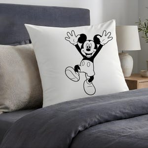 May include: White square pillow featuring a black and white illustration of Mickey Mouse with his arms raised in a joyful pose. The pillow is placed on a bed with gray bedding, creating a simple, clean aesthetic. A lamp and side table are visible in the background.
