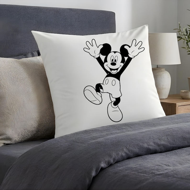 May include: White square pillow featuring a black and white illustration of Mickey Mouse with his arms raised in a joyful pose. The pillow is placed on a bed with gray bedding, creating a simple, clean aesthetic. A lamp and side table are visible in the background.