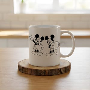 May include: White ceramic mug featuring a black and white illustration of Mickey Mouse and another character, each holding a heart. The mug sits on a round wooden coaster, with a white handle on the right side.