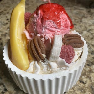 May include: A decorative candle shaped like a sundae, featuring a yellow banana, pink ice cream, red cherry, pecans, and white whipped cream in a white, fluted bowl. The candle has a wick.