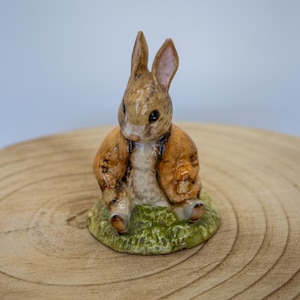 Może przedstawiać: Ceramiczna figurka siedzącego królika w brązowej kurtce. Królik ma brązowe i beżowe futro, czarne oczy i siedzi na kawałku zielonej trawy. Figurka stoi na drewnianej powierzchni.