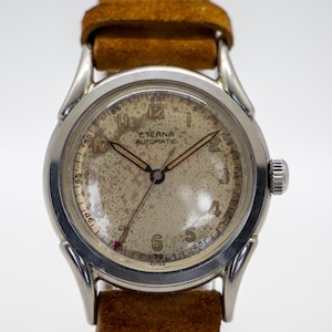 May include: Silver-toned Eterna Automatic wristwatch with a light-colored speckled face and brown leather band. The watch has a round face with black numerals and hands. The band is a light brown suede-like material.
