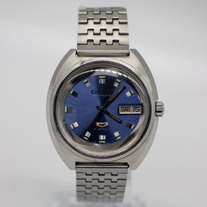 Pode incluir: Um relógio de pulso automático Citizen vintage com caixa e pulseira prateadas. O relógio apresenta um mostrador azul com marcadores prateados, um visor de dia e data e o texto "21 JEWELS". A pulseira do relógio é feita de elos de metal interligados.