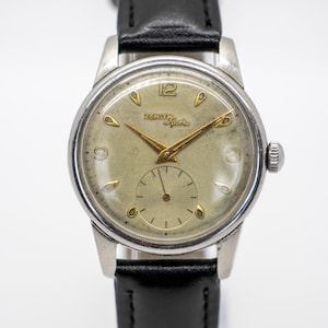 May include: A vintage Zenith Sporto wristwatch with a silver case and a black leather strap. The watch face is a light beige colour with gold hands and hour markers. It features a small seconds dial and the text "Zenith Sporto".