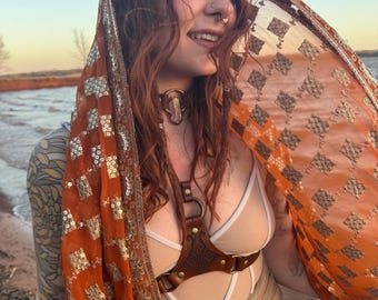 May include: A brown leather harness with decorative stitching and metal hardware. The harness is worn with a beige bodysuit. An orange and gold patterned scarf is draped over the shoulder.