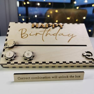 Puede incluir: Una caja de rompecabezas de cumpleaños de madera con las palabras "HAPPY Birthday" en letras doradas. La caja tiene una ranura, dos diales numéricos y un recorte en forma de llave. Un pequeño letrero de madera dice "La combinación correcta desbloqueará la caja."